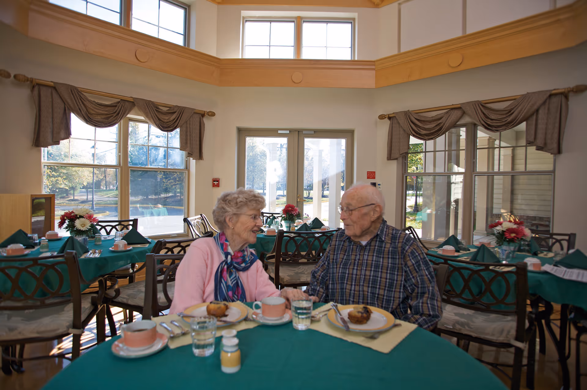 An elderly woman and man sitting at a round dining table in a bright room with large windows. The table is set with green tablecloths, plates with muffins, cups, glasses of water, and silverware. The woman is wearing a pink sweater and colorful scarf, and the man is wearing a plaid shirt. They are smiling and holding hands.