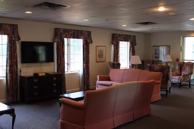 A cozy living room area with two pink upholstered sofas facing a flat-screen TV mounted on the wall. The room has multiple windows with patterned curtains, framed artwork on the walls, and several armchairs with floral upholstery. There are also table lamps on side tables, and the ceiling has recessed lighting.