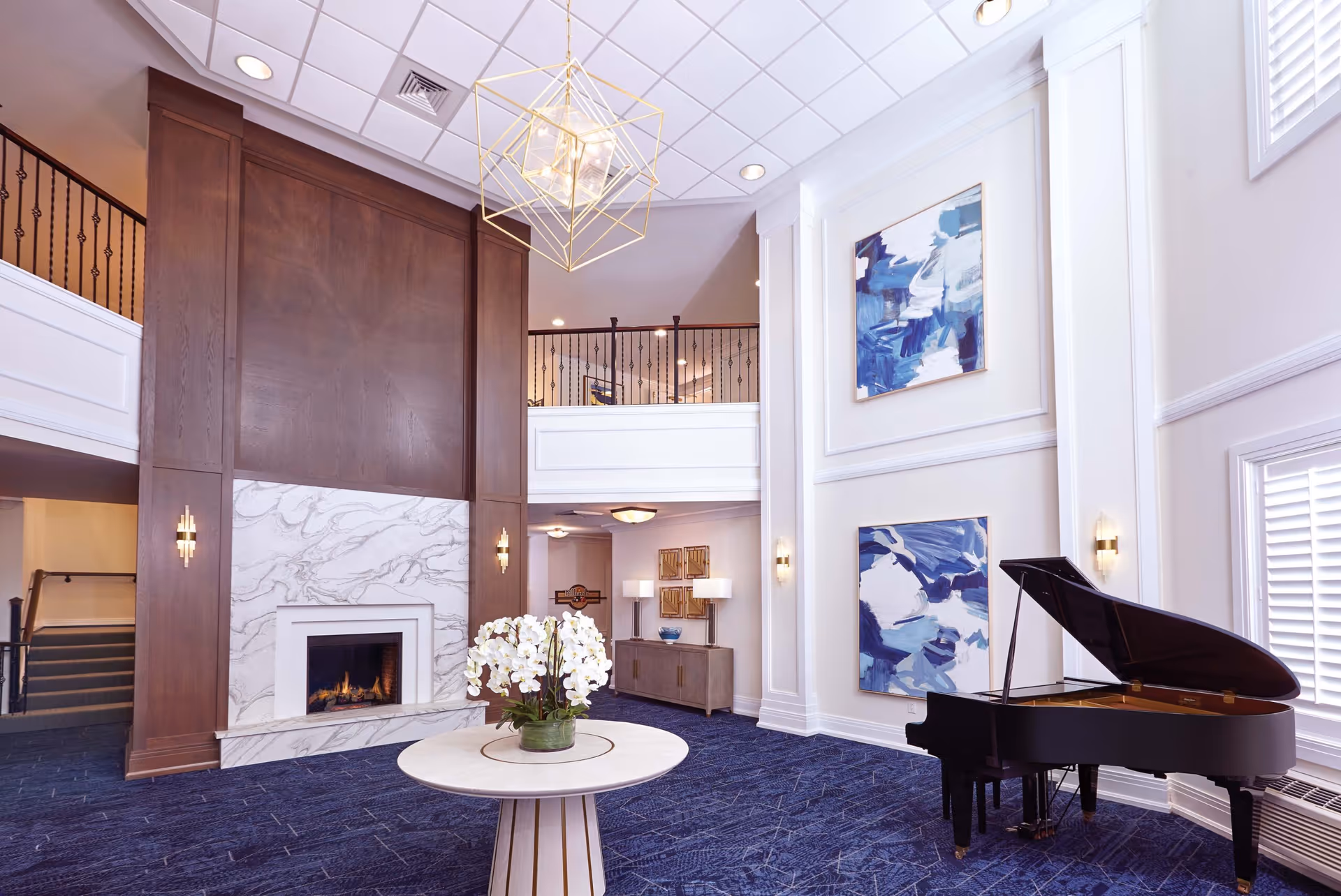 Two-story assisted living lobby with a fireplace, grand piano, a round table with flowers, and abstract wall art.