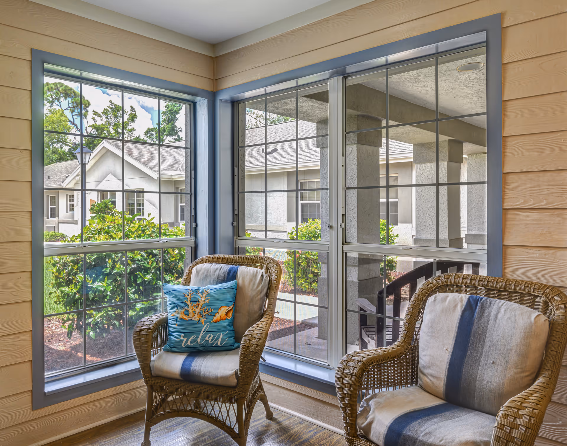 A cozy indoor seating area with two wicker chairs, each with cushions. One cushion has a blue pillow with seashell designs and the word 'relax'. Large windows with blue-gray frames provide a view of the outside greenery and neighboring buildings.