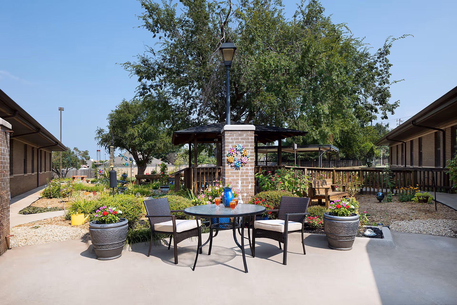 Sunny courtyard with a round table and two chairs surrounded by potted flowers, garden beds, a brick pillar, and a shaded gazebo between two single-story buildings.