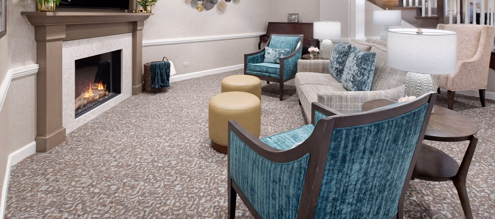 A cozy living room area with a lit fireplace, a beige patterned carpet, a beige sofa with blue patterned pillows, two blue upholstered armchairs, two round beige ottomans, a wooden side table with a lamp, and a piano in the background.
