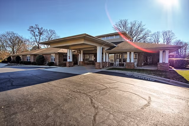 Exterior view of a single-story brick building with a covered entrance supported by white columns. The building is surrounded by leafless trees under a clear blue sky, and there is a paved driveway and parking area in front.