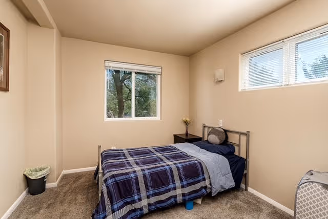 A small bedroom with beige walls and carpeted floor featuring a single bed with a blue and gray plaid comforter and a gray pillow. There are two windows, one on the left wall showing trees outside and a smaller one on the right wall. A small dark nightstand with a vase of flowers is next to the bed, and a trash bin is in the corner.
