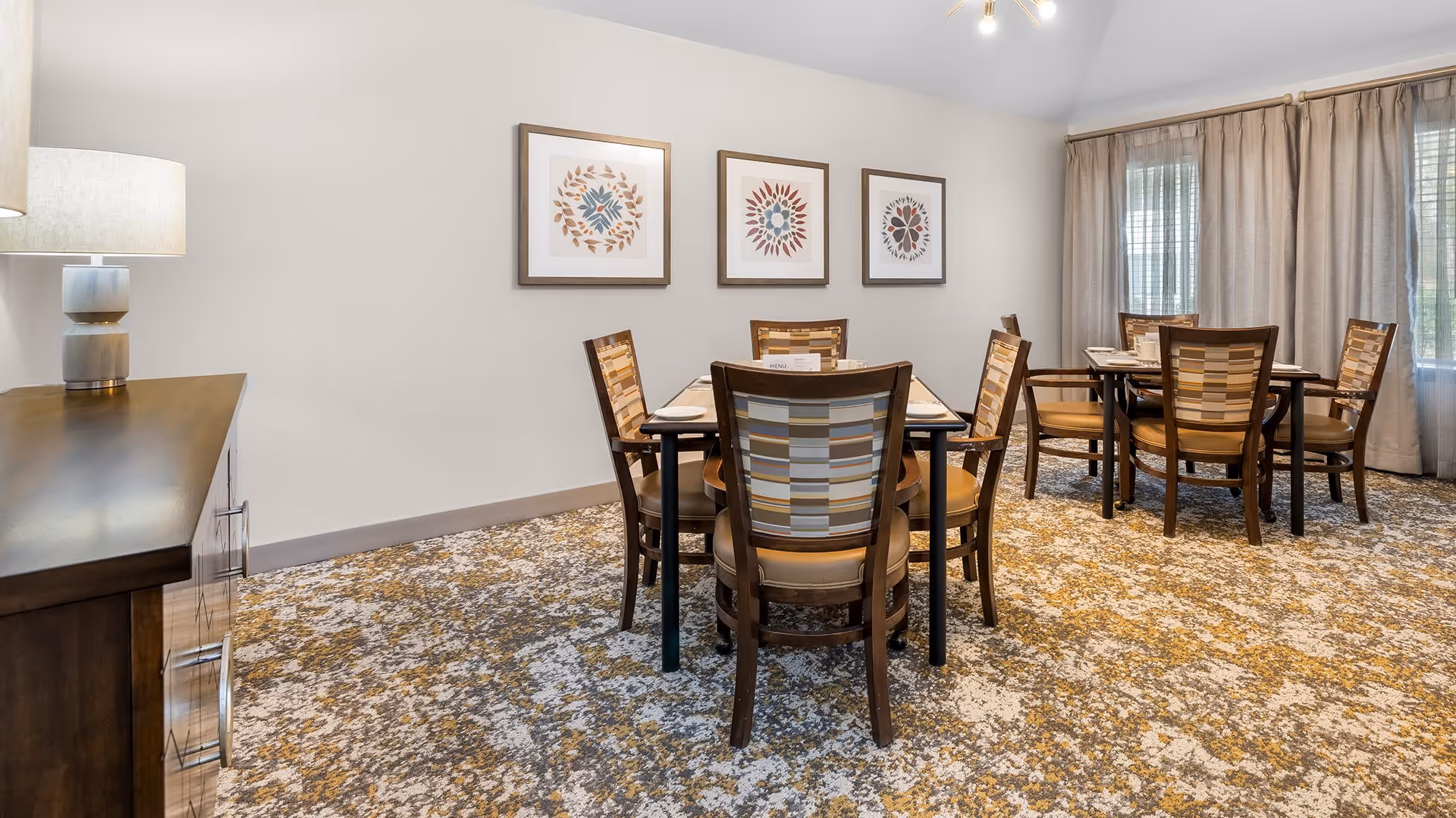 A dining room with two square tables, each surrounded by four wooden chairs with patterned backs and cushioned seats. The room has a patterned carpet in shades of beige, brown, and white. On the left side, there is a wooden sideboard with a lamp on top. Three framed abstract floral artworks hang on the wall above the tables. Large windows with beige curtains allow natural light into the room.