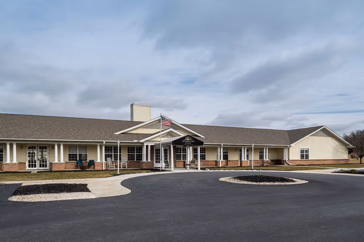 Exterior view of Celebration Villa Of Shippensburg, a single-story building with a covered entrance, an American flag on a flagpole, and a circular driveway in front. The building features beige siding with brick accents and multiple windows.