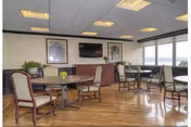 A bright and spacious common area with wooden flooring, several round tables with chairs, large windows letting in natural light, a wall-mounted television, framed artwork, and potted plants.