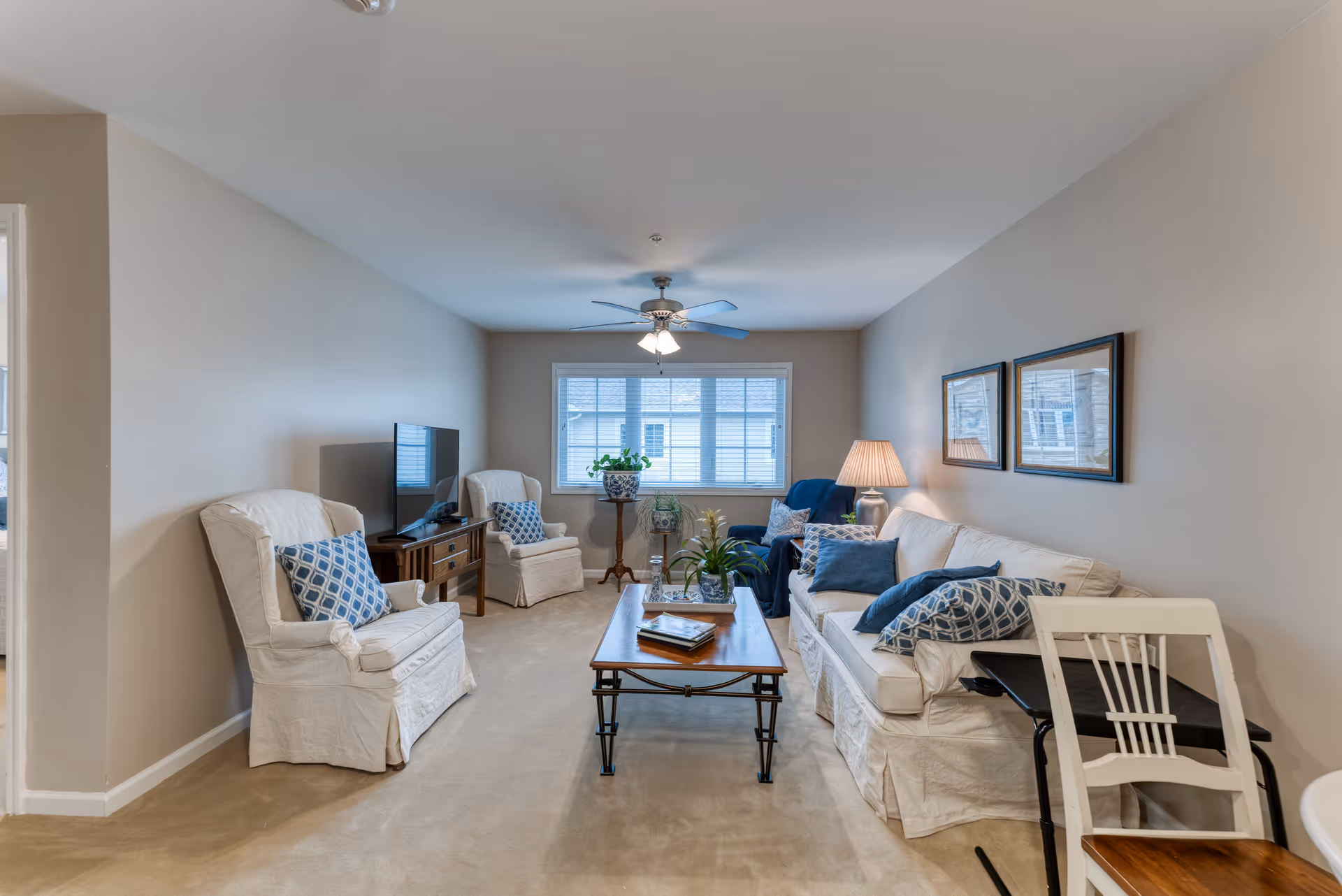 A cozy living room in a senior living facility featuring a white sofa with blue and patterned cushions, two white armchairs with patterned pillows, a wooden coffee table with books and a plant, a TV on a wooden stand, a ceiling fan with lights, a window with blinds, a table lamp, and framed pictures on the wall.