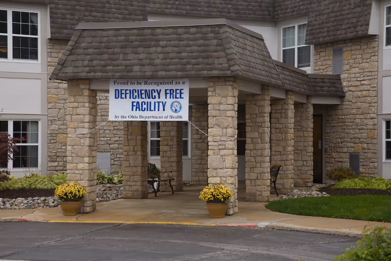 Stone-pillared entrance of a senior living building with a 'Deficiency Free Facility' banner and potted yellow flowers.