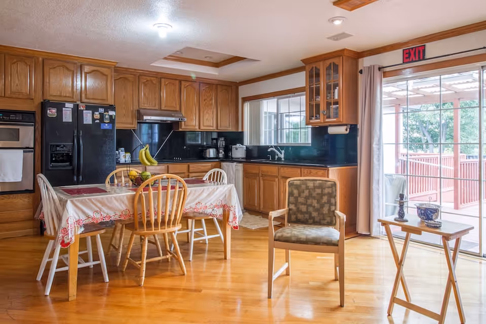 A kitchen and dining area with wooden cabinets, a black refrigerator, oven, and countertop appliances. A dining table covered with a white and red tablecloth is surrounded by mismatched chairs. A single cushioned chair and a small wooden side table with decorative items are near a sliding glass door leading to an outdoor patio with a red railing.