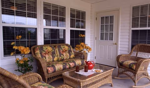 A cozy enclosed porch area with wicker furniture including a loveseat, two chairs, and a coffee table. The loveseat and chairs have floral patterned cushions. On the coffee table, there is a red pitcher, two glasses, and a plate with snacks. The porch has white walls, multiple windows with blinds, and a white door with a window.