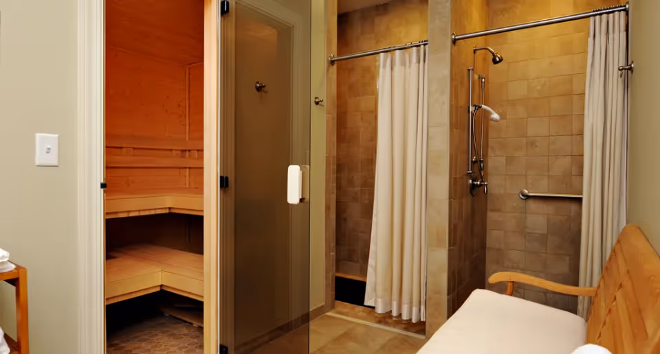 Interior view of a wellness area featuring a wooden sauna room with benches on the left, two tiled shower stalls with white curtains in the center and right, and a wooden bench with a white cushion on the right side.