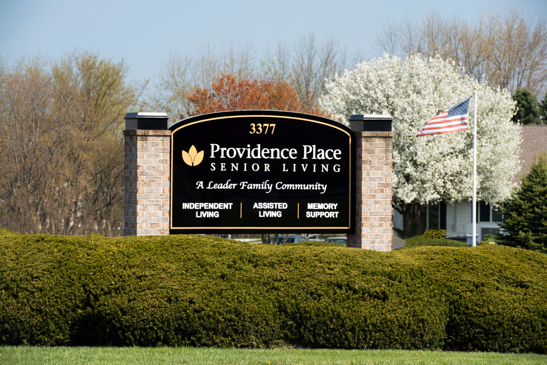 Outdoor view of a large black and gold sign for Providence Place Senior Living of Dover, surrounded by green bushes with trees and an American flag in the background.