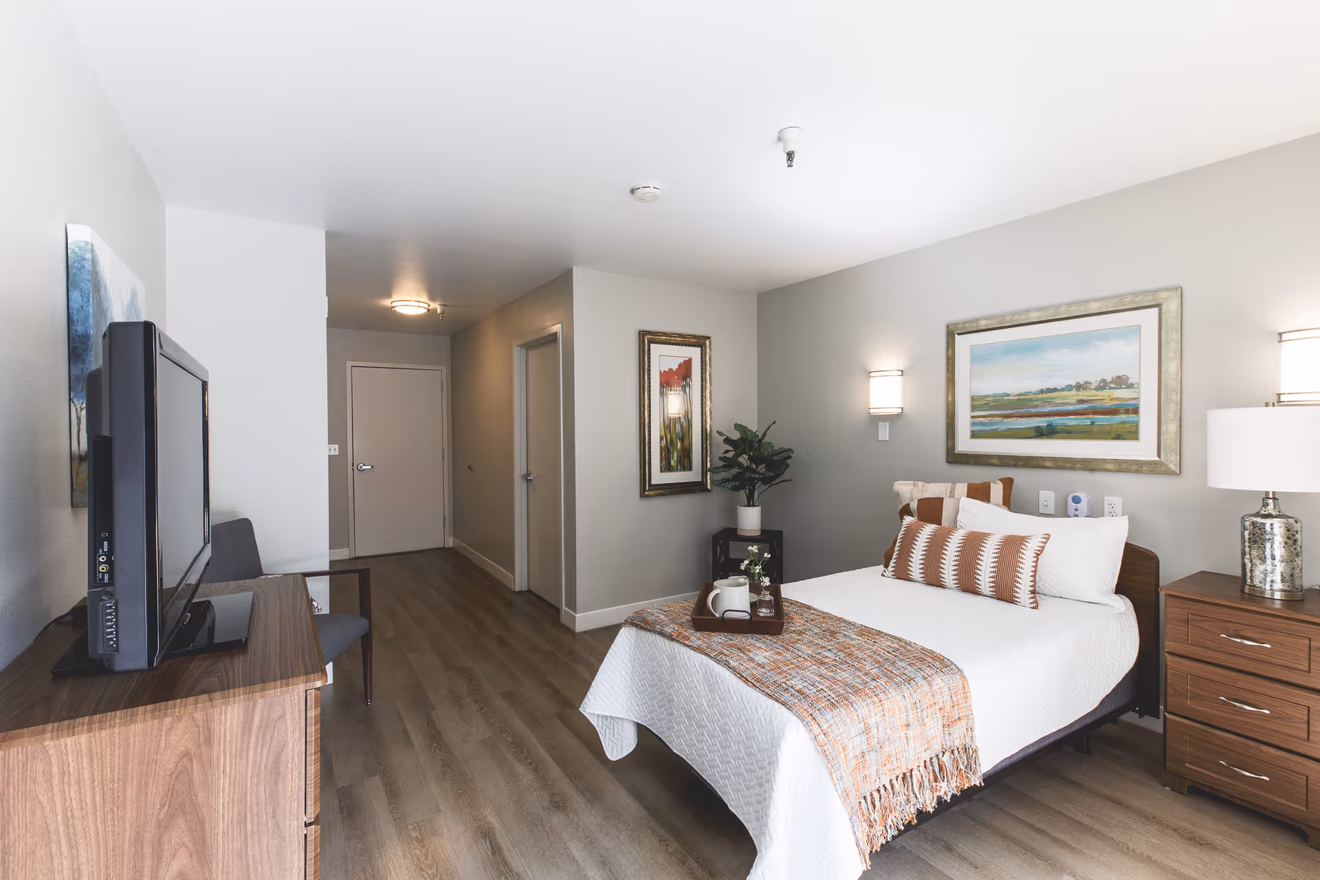 A neatly arranged bedroom in a senior living facility featuring a single bed with white bedding and a decorative throw and pillows. There is a wooden nightstand with a lamp on the right side of the bed, a framed landscape painting above the bed, and a small plant on a black stand in the corner. A flat-screen TV is placed on a wooden dresser opposite the bed, with a chair next to it. The room has light-colored walls and wood flooring, with two doors visible in the background.