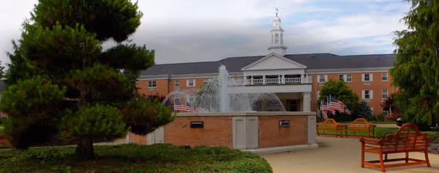 Front exterior of Copeland Oaks showing a brick building with a central cupola, a fountain, benches, American flags, and landscaped grounds.