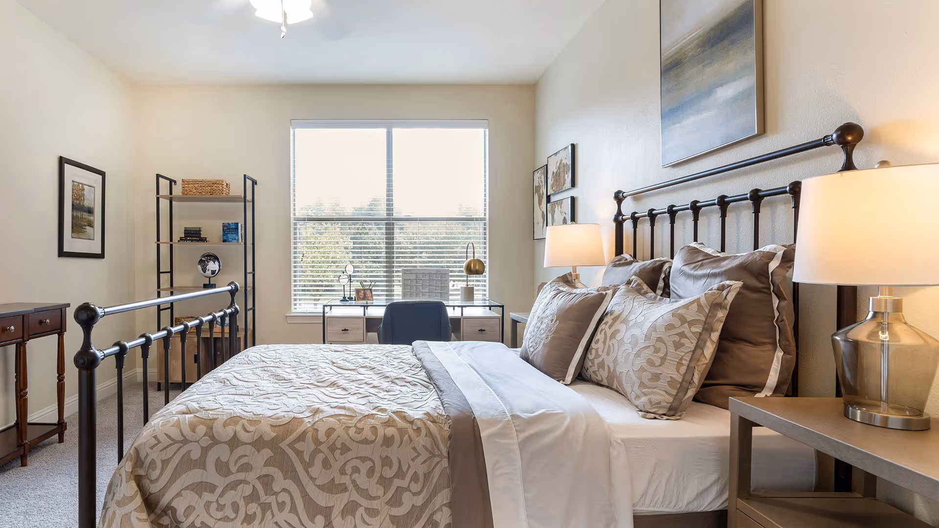 A bright bedroom with a large window letting in natural light. The room features a metal bed frame with beige and patterned bedding, two bedside tables each with a lamp, a desk with a chair in front of the window, a shelving unit with decorative items, and framed artwork on the walls.