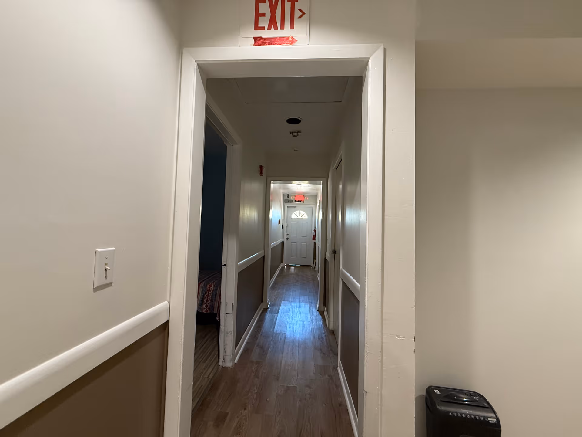A narrow hallway with wooden flooring and beige walls, featuring several doorways along the sides. An exit sign is mounted above the doorway at the near end of the hallway, and a white door with a window is visible at the far end. A small black appliance is positioned against the right wall near the foreground.