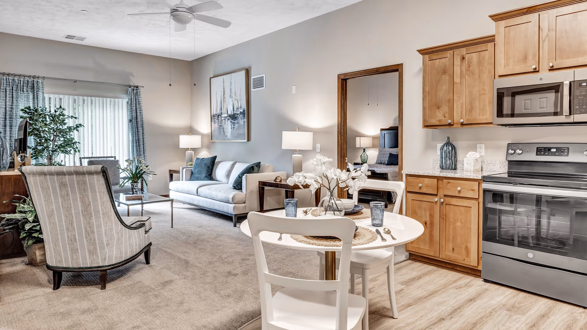 A bright and cozy living space in a senior living facility featuring a small round dining table set for two with white chairs and decorative flowers. The room includes a comfortable sofa with cushions, an armchair, a coffee table, and a TV on a wooden stand. The kitchen area has wooden cabinets, a stainless steel oven, and a microwave. A large mirror on the wall reflects a bedroom with a bed and lamps. The room is carpeted with light-colored flooring in the kitchen area and has a ceiling fan and large window with curtains.