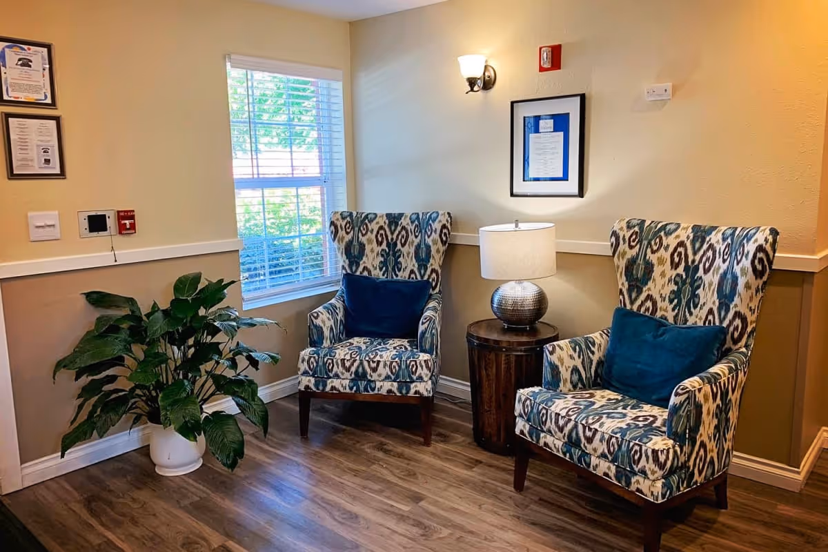 A cozy corner in a senior living facility with two patterned armchairs featuring blue and beige designs, each with a blue cushion. Between the chairs is a round wooden side table with a silver textured lamp. A large green potted plant sits near a window with white blinds, and framed documents hang on the beige walls. The floor is wood with a warm tone.
