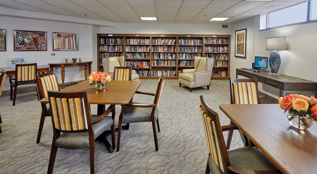 A cozy senior living library and lounge area with bookshelves filled with books along the back wall, two beige armchairs, several wooden tables with striped cushioned chairs, and vases of flowers on the tables. There is a desk with a computer and a lamp on the right side, and framed artwork on the walls.