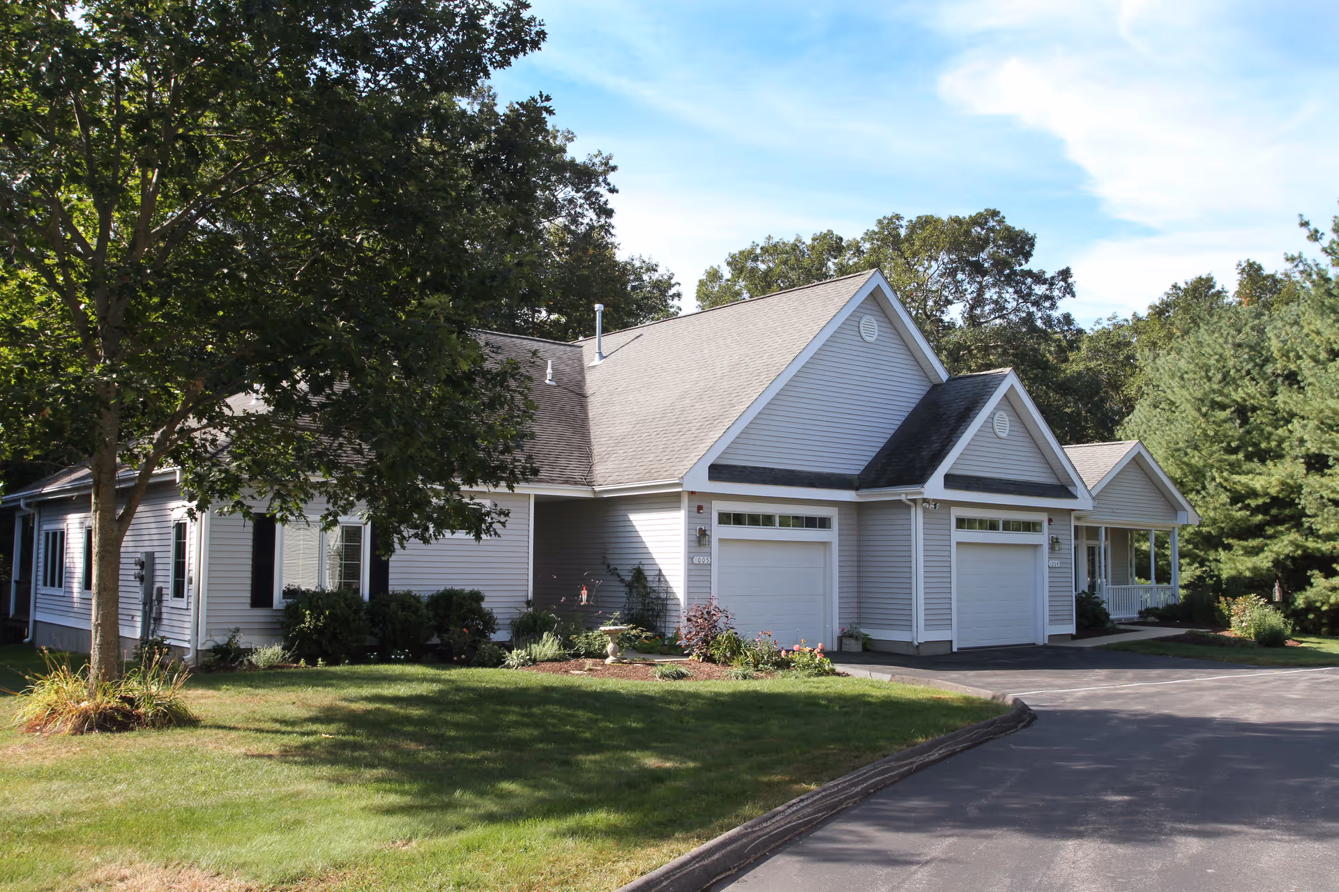 Exterior view of a single-story residential building with light gray siding, white trim, and two garage doors. The building is surrounded by green grass, trees, and shrubs under a clear blue sky.