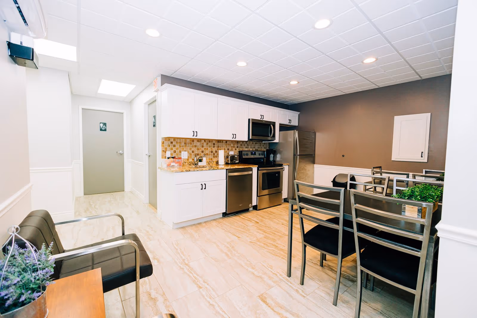A modern kitchen and dining area with white cabinets, stainless steel appliances including a refrigerator, stove, microwave, and dishwasher. There is a dining table with metal chairs and a small seating area with a black cushioned chair and a small wooden table with a potted plant. The floor is tiled, and the walls are painted white and gray. Two doors are visible in the background, one with a restroom sign.