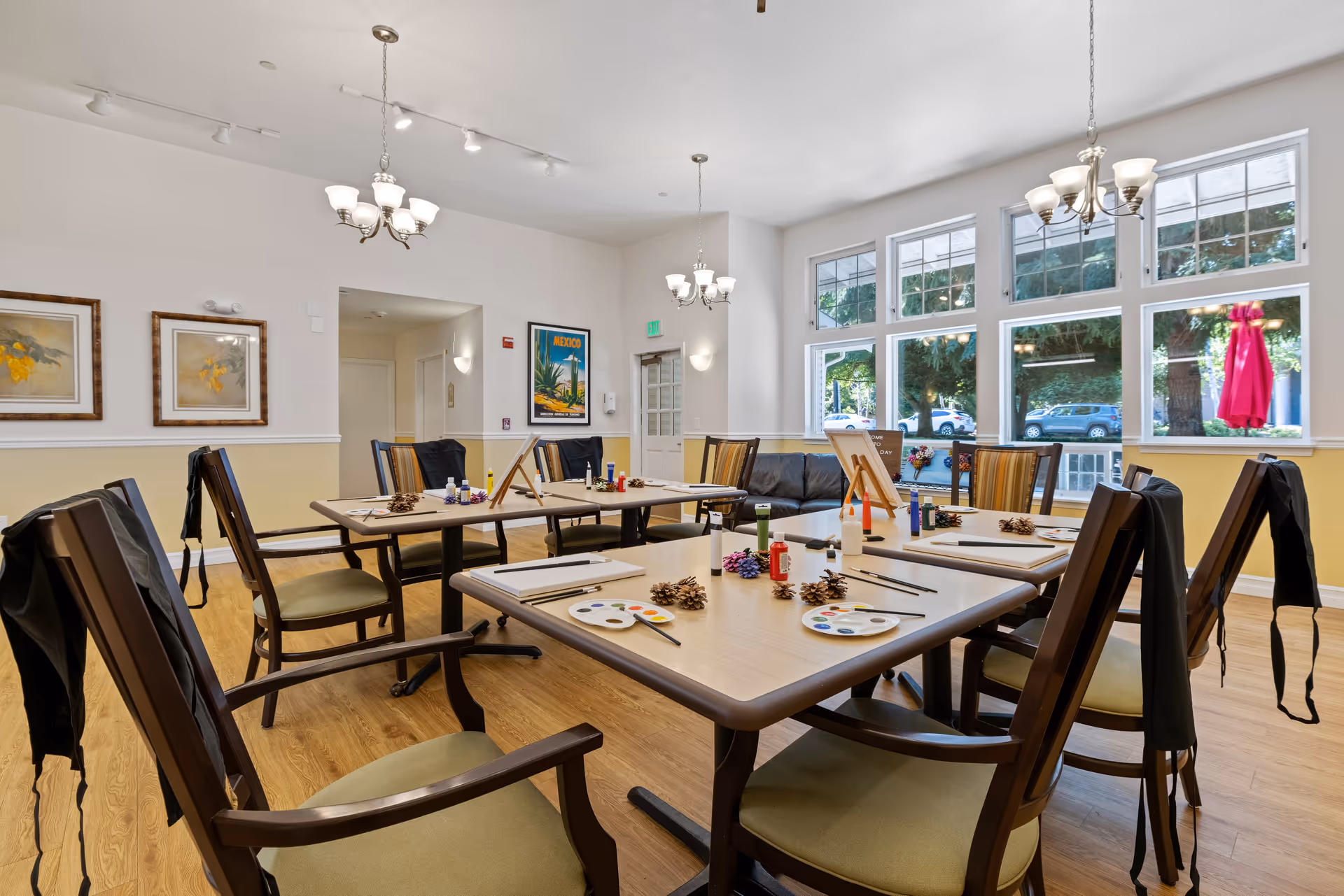 A bright and spacious activity room with large windows letting in natural light. Several tables are arranged with painting supplies including paint palettes, brushes, and canvases on easels. Chairs with aprons draped over the backs surround the tables. The room has light-colored walls, wooden flooring, and decorative framed artwork on the walls.