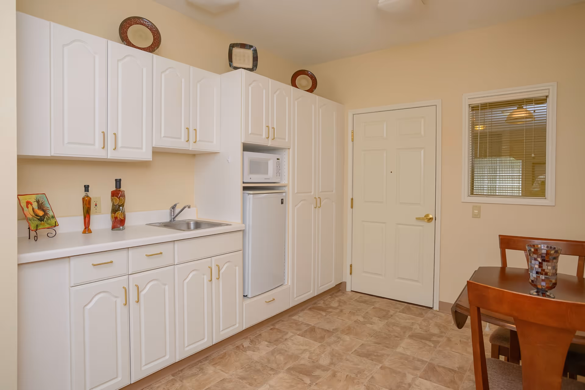 Interior view of a small kitchen area with white cabinets, a countertop with a sink, a microwave, and a mini refrigerator. There are decorative plates on top of the cabinets and two decorative bottles on the counter. To the right, there is a white door and a window with blinds, and part of a wooden dining table with chairs is visible.