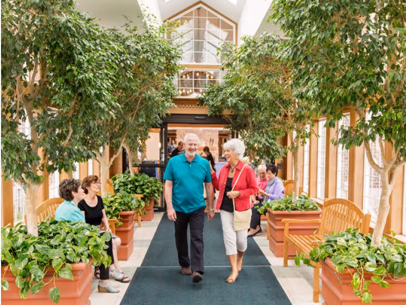 A bright indoor hallway in a senior living facility with large potted plants and wooden benches along the sides. Several elderly people are sitting and chatting on the benches, while an elderly man and woman walk hand in hand down the center carpeted walkway, smiling and enjoying the space.