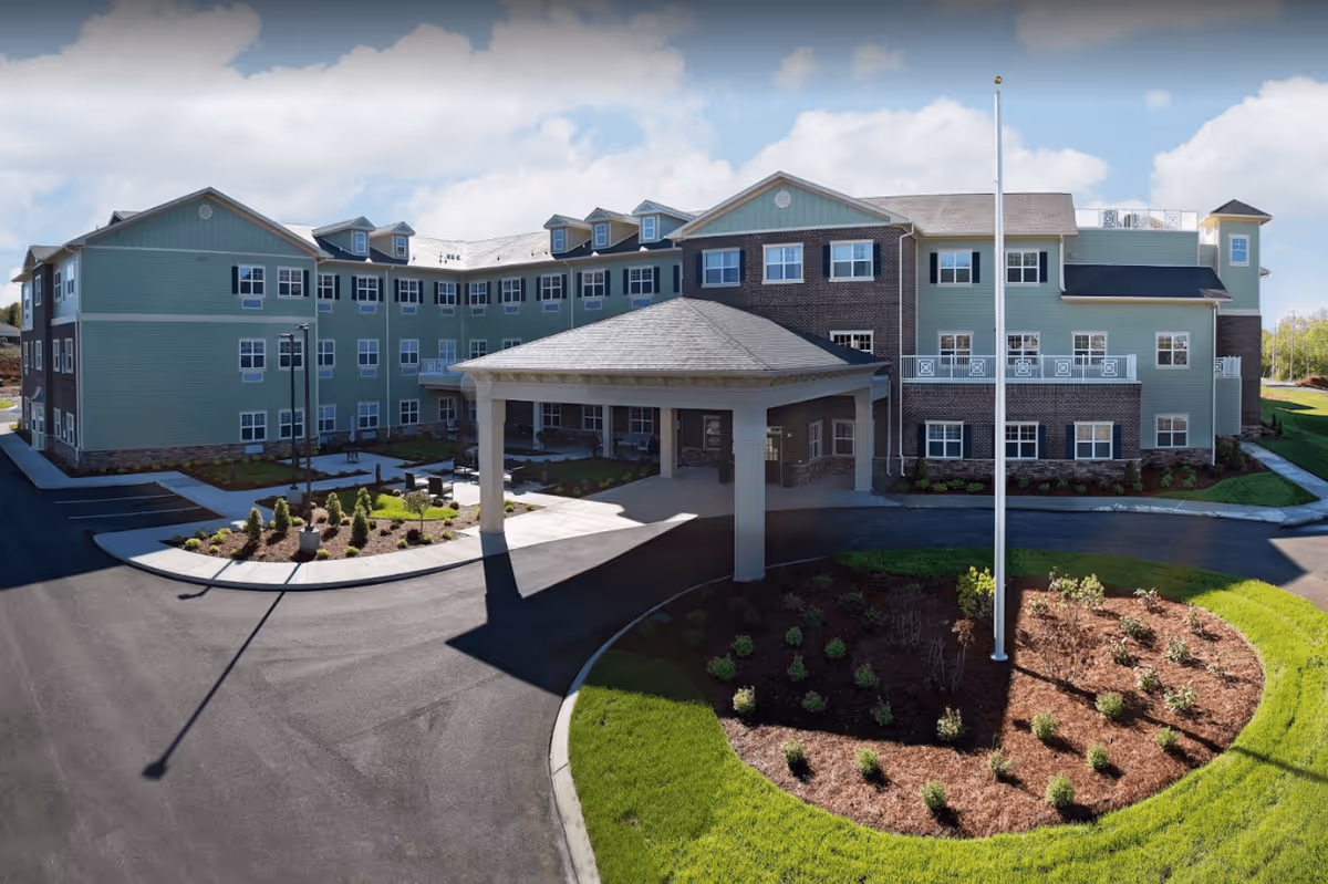 Exterior view of a large, three-story senior living facility building with green siding and brick accents. The building has multiple windows, a covered entrance with columns, a circular driveway, landscaped garden beds, and a flagpole in front. The sky is partly cloudy.