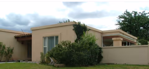 Single-story stucco building with a low courtyard wall, shrubs and grass under a cloudy sky.