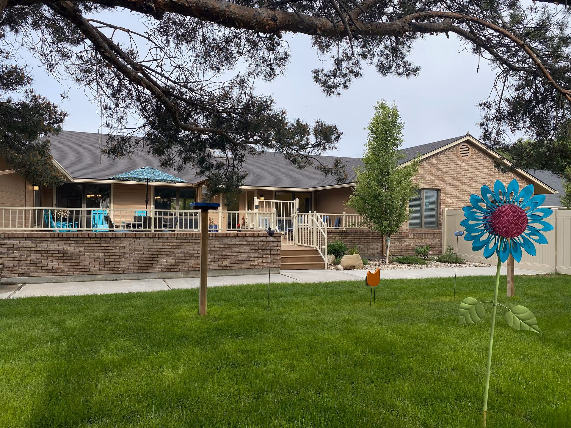 Outdoor view of a single-story brick and siding building with a patio area featuring turquoise chairs and an umbrella. The foreground shows a green lawn with decorative garden stakes shaped like flowers and a bird feeder. Trees frame the top of the image.