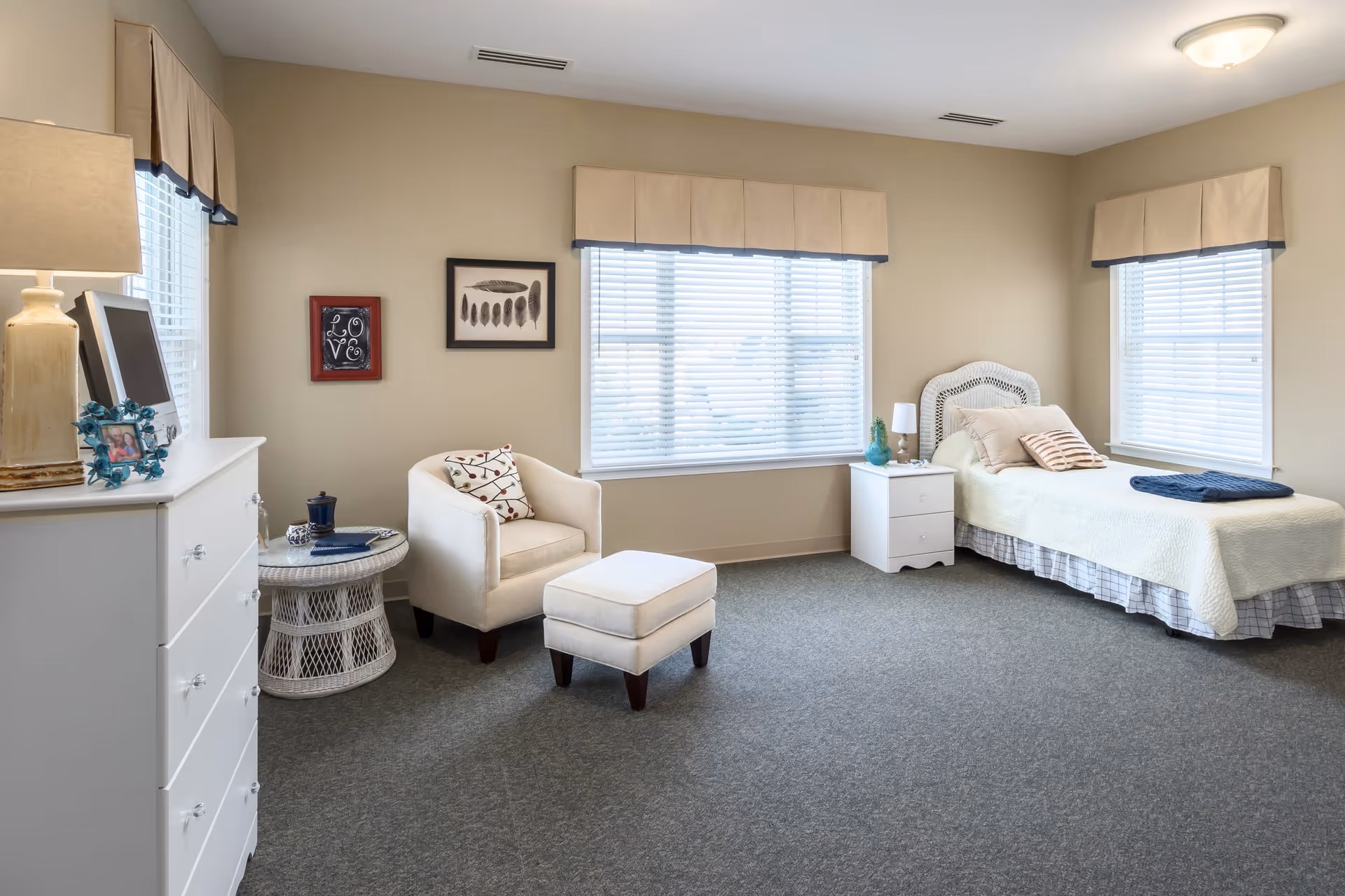 A bright and spacious bedroom with beige walls and carpeted floor. The room features a single bed with a white quilt and pillows, a white nightstand with a lamp and decorative items, a white armchair with a matching ottoman, a round wicker side table, a white dresser with a lamp and framed photo, and three windows with beige valances allowing natural light to fill the room. Two framed artworks hang on the wall above the armchair.