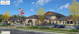 Exterior view of The Pavilion Senior Living at Lebanon, showing a single-story building with a stone facade and multiple windows. There are two flagpoles with the American flag and a state flag in front of the building. Several people are walking near the entrance, and there are parked cars in the parking lot. Trees with yellow leaves and landscaping surround the area under a clear blue sky.