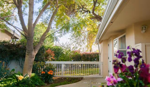 A peaceful outdoor garden area with a large tree, colorful flowers, and green shrubs next to a beige building with windows and a white fence in the background.