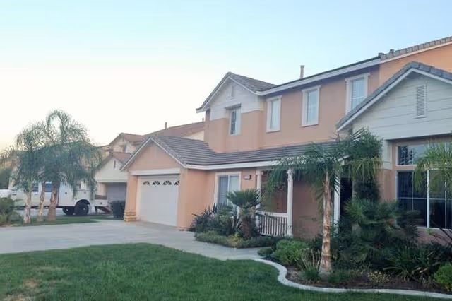 Exterior view of a two-story residential house with a beige and white facade, a garage, a driveway, and a well-maintained front yard with palm trees and shrubs under a clear sky.