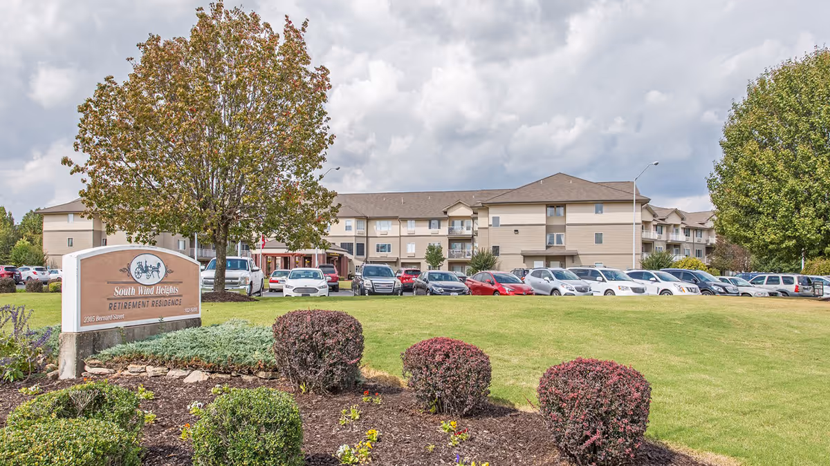Front exterior of South Wind Heights retirement residence with its entrance sign, landscaped lawn, parked cars, and a three-story building under a cloudy sky.
