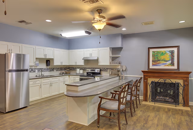 A spacious kitchen area with white cabinets, stainless steel refrigerator, stove, and a long countertop with three wooden chairs. The room has a ceiling fan with a light, wood flooring, and a decorative fireplace with a framed painting above it on the right wall.