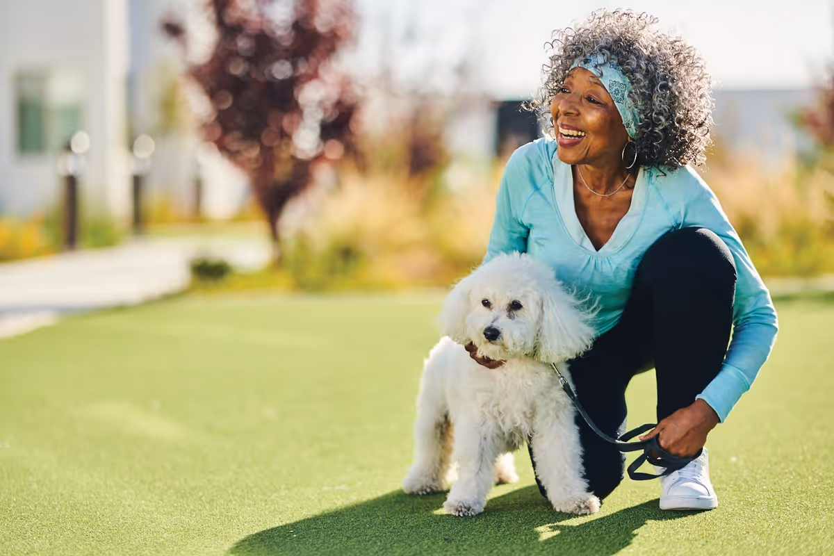 A joyful elderly woman with curly gray hair, wearing a light blue long-sleeve shirt and black pants, crouches on a grassy area while holding a small white fluffy dog on a leash. The background shows blurred outdoor greenery and buildings.