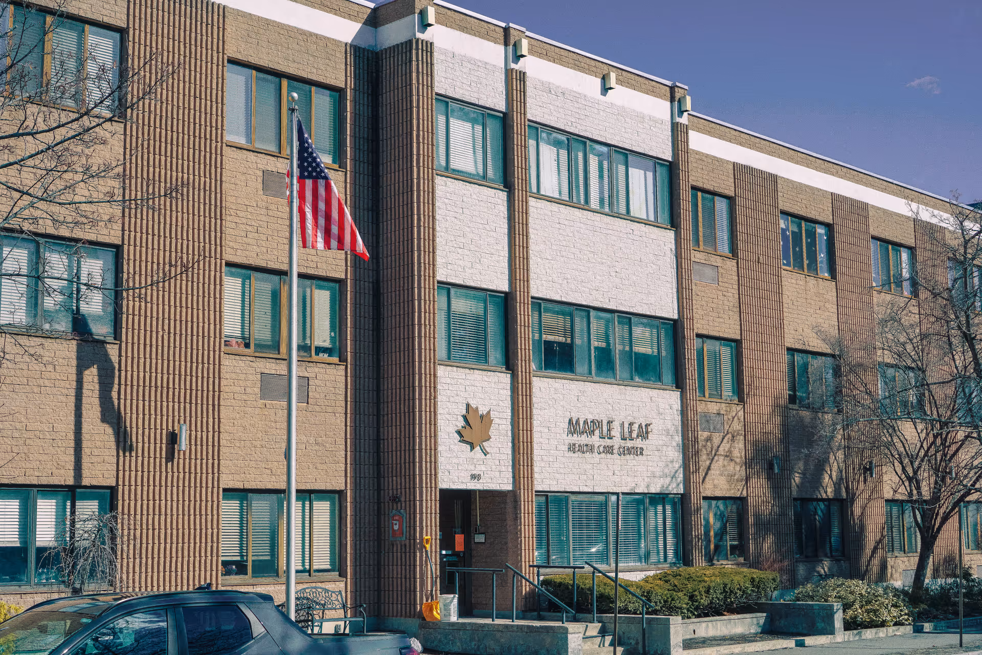 Front exterior of the Maple Leaf HealthCare Center building showing the entrance, windows, and an American flag.