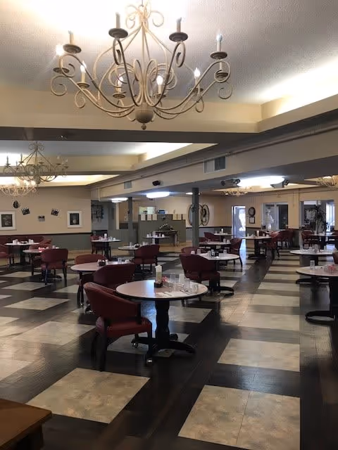 Spacious dining room with multiple round tables and red cushioned chairs arranged neatly. The floor has a checkered pattern with dark and light tiles. Elegant chandeliers hang from the ceiling, and the room is well-lit with natural and artificial light. Walls are decorated with framed pictures and there are large windows and doors visible in the background.