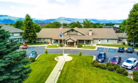 Front exterior view of Missoula Valley Senior & Assisted Living (The Auberge at Missoula Valley) building with a driveway, parking spaces, several cars, a flagpole with American and state flags, green lawns, trees, and mountains in the background under a partly cloudy sky.