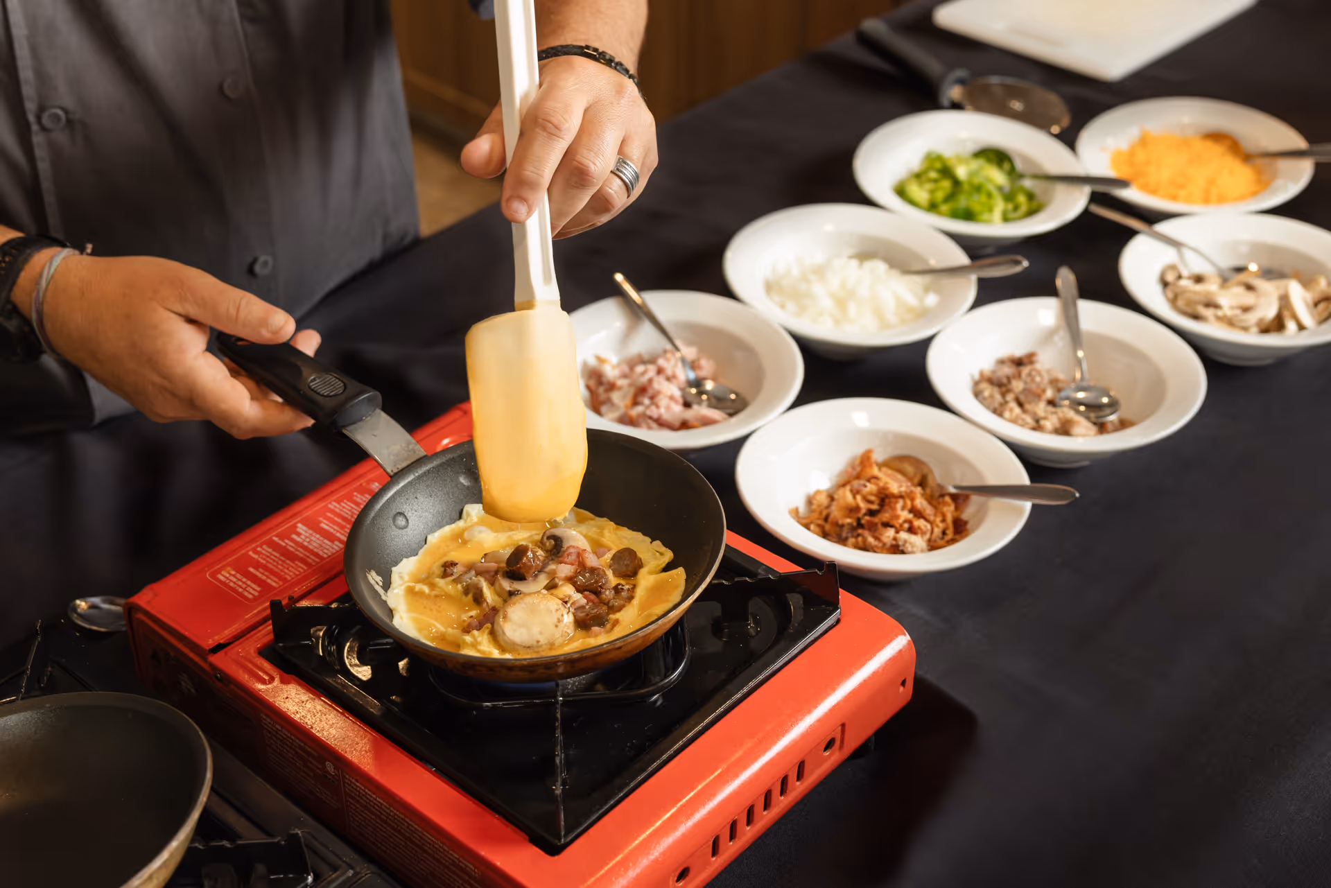 A person cooking an omelette in a frying pan on a portable gas stove, with several bowls of chopped ingredients like green peppers, onions, cheese, mushrooms, bacon, and sausage on a black tablecloth nearby.
