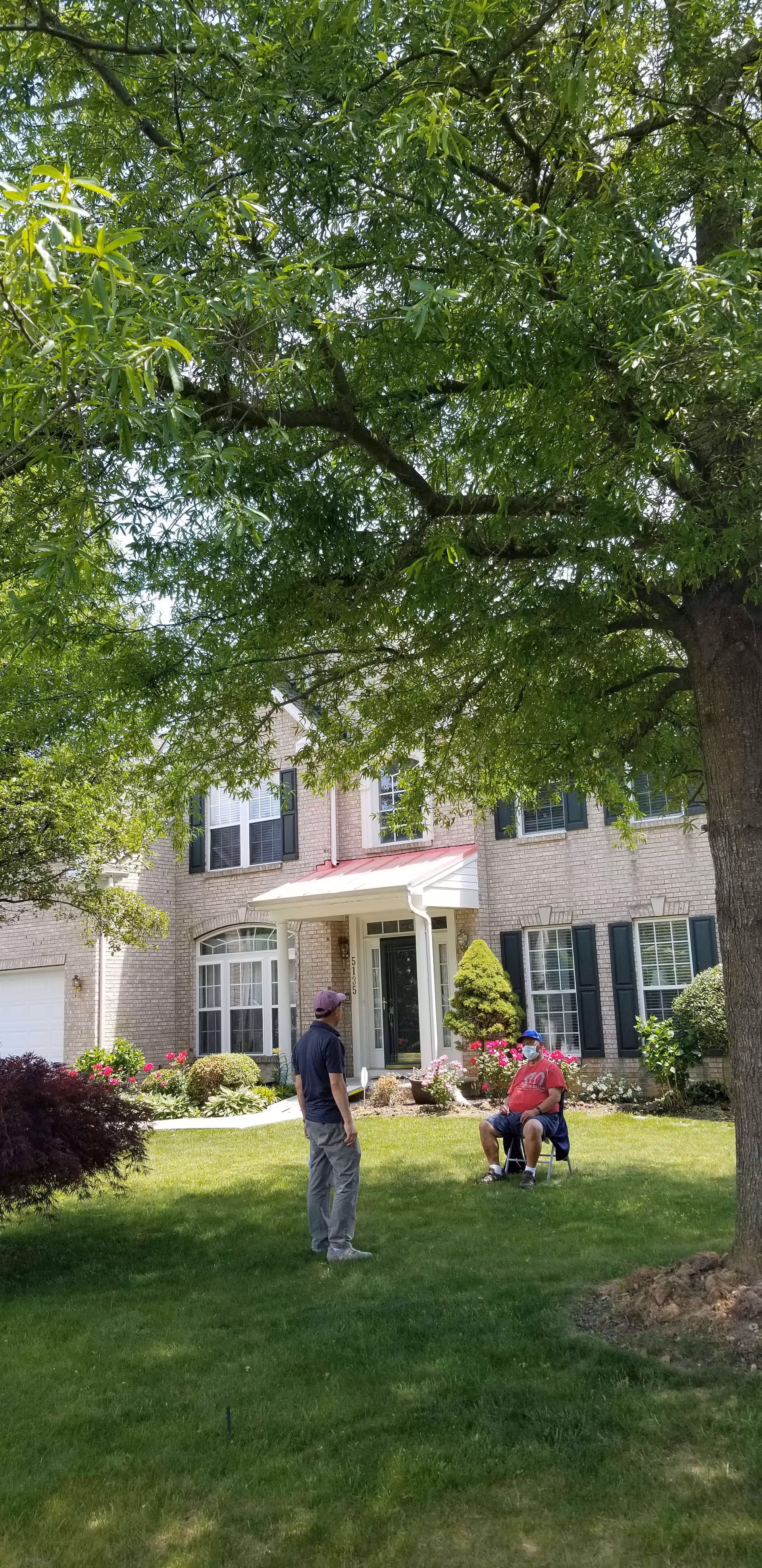 Two men in a grassy front yard of a large brick house with black shutters and a white porch. One man is standing and the other is sitting on a folding chair under a large tree with green leaves. The yard has bushes and flowers near the house.