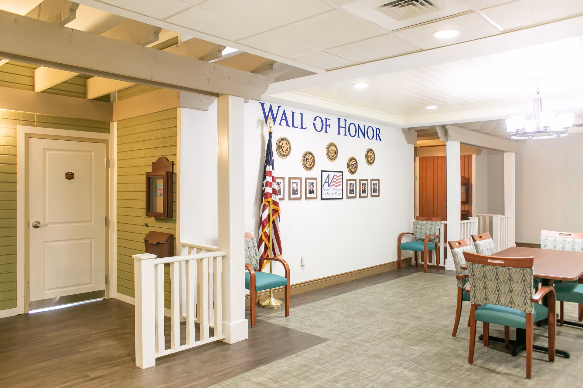Interior view of a senior living facility hallway featuring a Wall of Honor with framed photos and military emblems, an American flag, several chairs with teal cushions, a wooden table with chairs, and a white door on the left side.