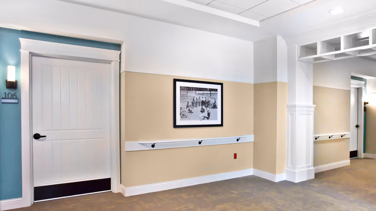 Interior hallway of a senior living facility with beige and white walls, a white door labeled 106, a black and white framed photograph on the wall, handrails along the walls, and ceiling lights.