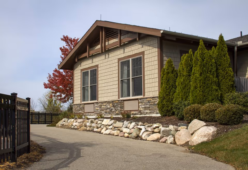Driveway alongside a single-story building with beige siding, stone foundation, and landscaped shrubs and rocks.