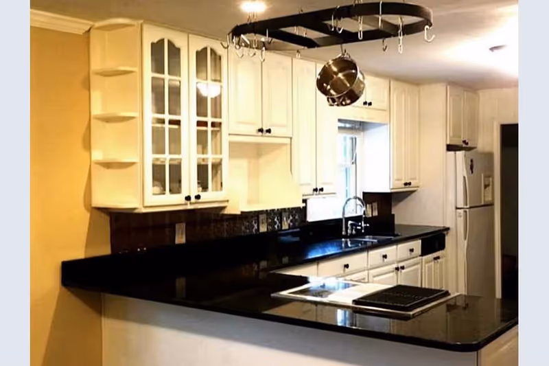 A modern kitchen with white cabinets and black countertops. There is a hanging pot rack with a single pot above the counter. The kitchen features a double sink under a window, a refrigerator, and a stove built into the counter. The walls are painted yellow and white.