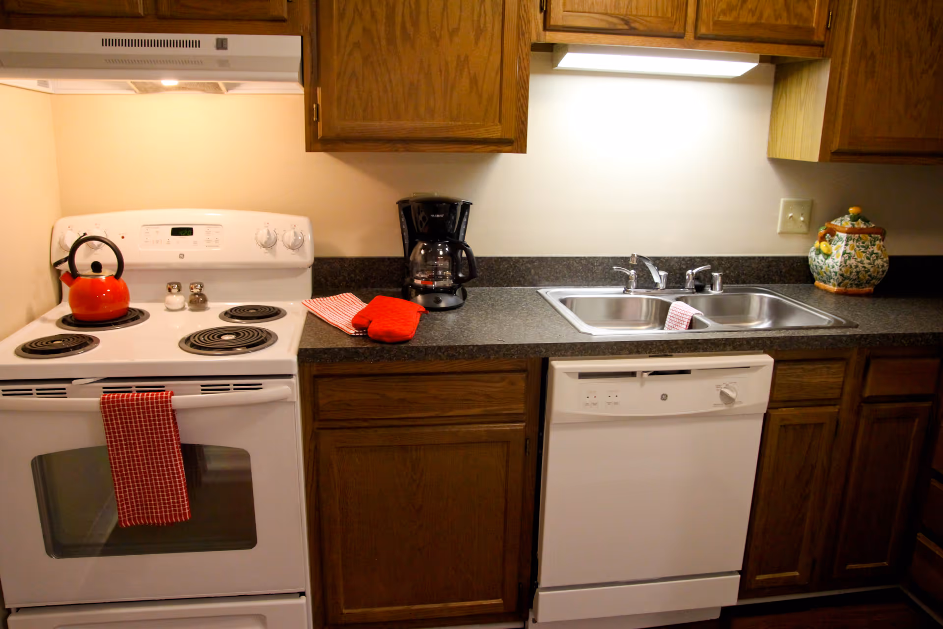 A kitchen area featuring a white electric stove with a red kettle on one burner and a red checkered towel hanging from the oven handle. Next to the stove is a countertop with a coffee maker, a red oven mitt, and a red and white striped cloth. There is a double stainless steel sink with a faucet, and a dishwasher below the counter. Wooden cabinets are above and below the countertop, and the area is well-lit with under-cabinet lighting.