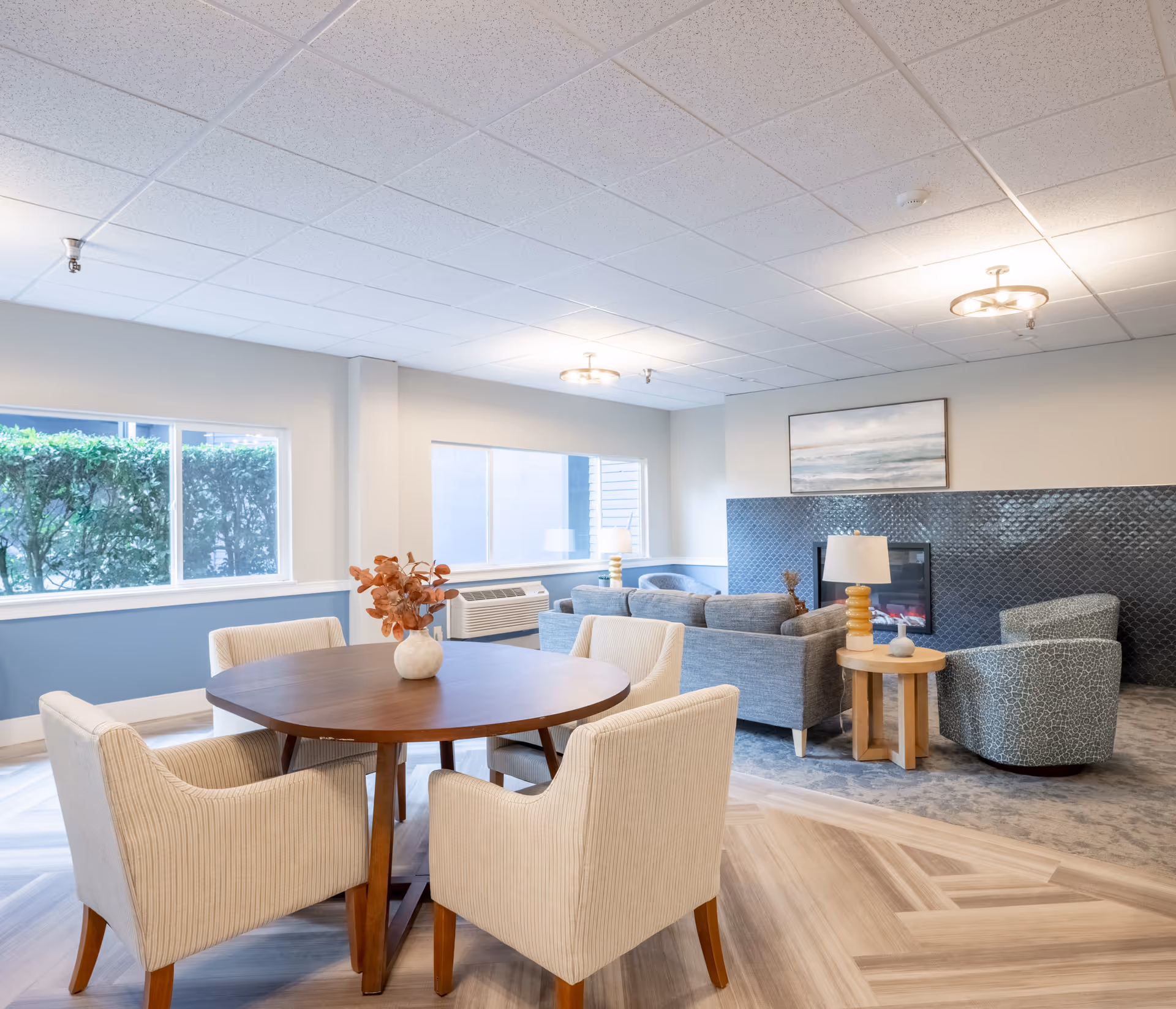 Bright senior living common room with a round wooden table and upholstered chairs in the foreground and sofas with a fireplace in the background.